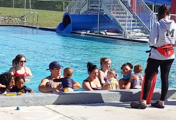 six parents with toddlers in outdoor pool. swim instructor standing on pool deck giving directions