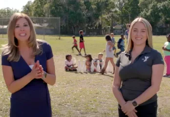 Still from video of Gayle Guyardo and Alyssa Heartstock outside in empty field, smiling. Kids playing games in after school program in the background.