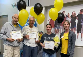 Three Greater Palm Harbor Y LIVESTRONG® at the YMCA participants and their coach at their graduation ceremony in April 2022. Balloons and poster of happy people side hugging in background.