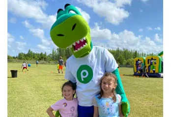 Two children at 30th Healthy Kids Day® pose for a photo with Publix mascot, Plato the Publixaurus. Background: kids playing sports and inflatable obstacle course.