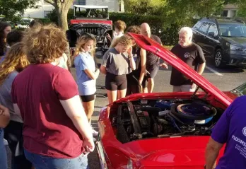 A group of teens from Teen Leaders Club at John Geigle gather around a red car with the hood lifted. Car aficionado and YMCA namesake John Geigle teaches the group about caring for cars. 