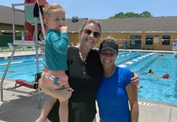 Swim instructor and mom holding young child on the outdoor pool deck. In the background, people are swimming and a lifeguard in a chair watches over them. 