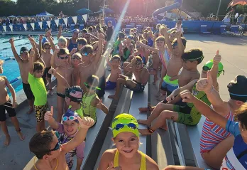 A group of 50+ youth triathletes smile while holding thumbs up on the outdoor pool deck.