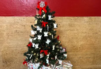 A small Christmas tree with paper angel ornaments and red bows in front of red plus tan stone wall.
