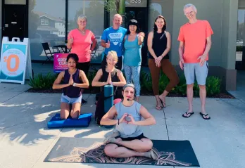YMCA staff and members smiling outside new facility. Three people are on the ground in yoga pose with prayer hands and five others stand behind them.