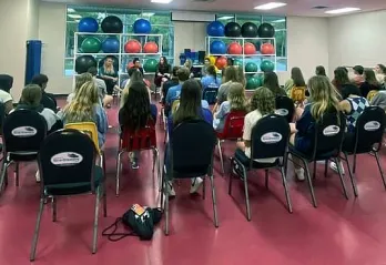 Teens in a large room with red floors, sitting in chairs listening to adult speaker during a meeting.