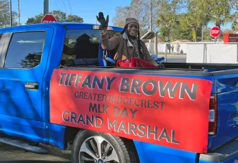 Ridgecrest leader waves from the bed of a blue truck for Greater Ridgecrest MLK Day parade. Sign is on side of truck bed, black block letters in all caps: Tiffany Brown Greater Ridgecrest MLK Grand Marshal on red background.