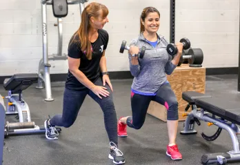 Female personal trainer demonstrates a forward lunge to female client, as client is in the stance. Client is holding dumbbells in bicep curl position.