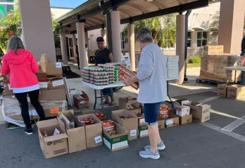 Four volunteers at James P. Gills YMCA organize food donations outside in the shade. Many brown boxes of food rest on pallets and the ground. Foreground includes a man facing away from the camera, holding cereal boxes.