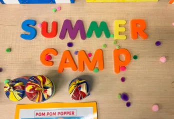 Camp craft call pom pom popper shot from above. Child play letters spell summer camp. Pom poms scattered on the table scene.