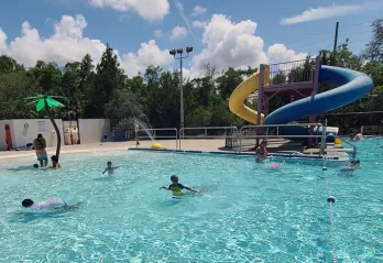 Kids playing in outdoor zero-entry pool with waterslides in the background. Sunny day with puffy white clouds and tall evergreen trees in background scenery.