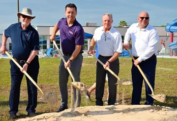 Outdoor image of four men posing for a groundbreaking photo. The subjects are holding shovels with dirt. A swimming pool with slides is in the background.
