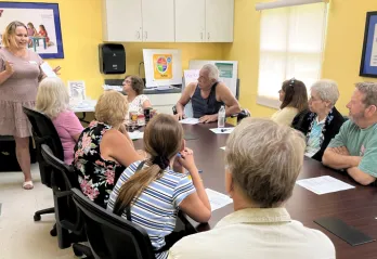 A group of nine adults sitting at a table, listening to a woman standing, teaching the class. Background: classroom walls are yellow.