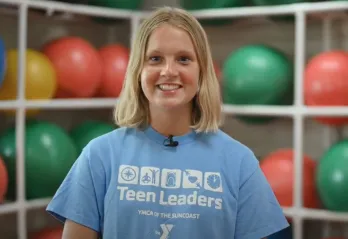 A teen girl with short blonde hair and a light blue Teen Leaders YMCA shirt smile at the camera. The background is out of focus, and shows white shelves holding green and red exercise balls.
