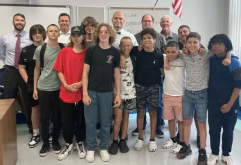 A group of five adults stand behind 10 middle school students, posing for a photo. The setting is a school classroom, a portion of the hanging American flag is shown in the background.