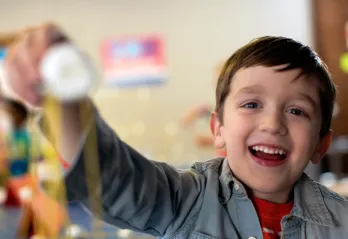 A young boy smiles as he reaches his arm out to show off the craft he made. The child's face is in focus and the craft is in the foreground, out of focus.