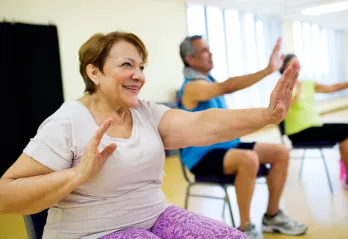 Three seniors holding arms out during chair exercises in fall prevention program. Woman wearing purple pants is in focus, two seniors in the background are out of focus.