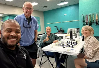 4 members of Chess Club smiling for photo. Two are sitting beside the chess table.