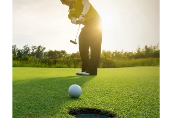 Foreground: golf ball rolling into hole. Background: man in golf attire putting at sunrise. Trees behind him.