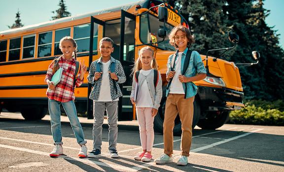 4 children smiling outside school bus with backpacks