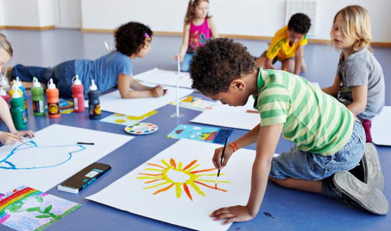 A group of six school age children set up with painting supplies and canvases on the floor painting.