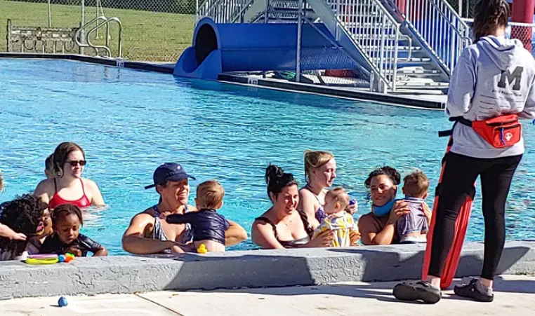 six parents with toddlers in outdoor pool. swim instructor standing on pool deck giving directions