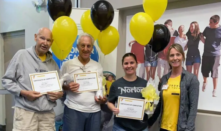 Three Greater Palm Harbor Y LIVESTRONG® at the YMCA participants and their coach at their graduation ceremony in April 2022. Balloons and poster of happy people side hugging in background.