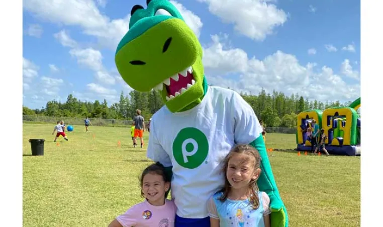 Two children at 30th Healthy Kids Day® pose for a photo with Publix mascot, Plato the Publixaurus. Background: kids playing sports and inflatable obstacle course.