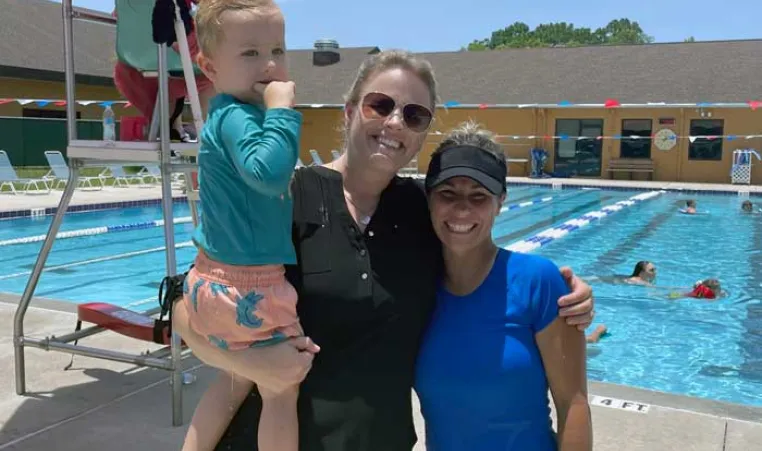 Swim instructor and mom holding young child on the outdoor pool deck. In the background, people are swimming and a lifeguard in a chair watches over them. 