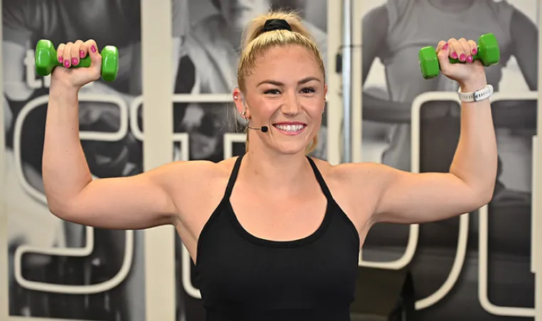 Exercise instructor smiling and flexing muscles while holding weight.s