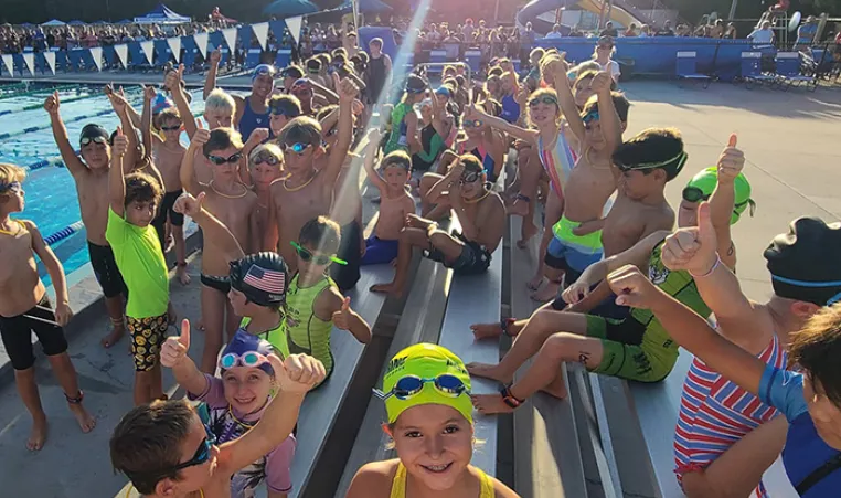 A group of 50+ youth triathletes smile while holding thumbs up on the outdoor pool deck.