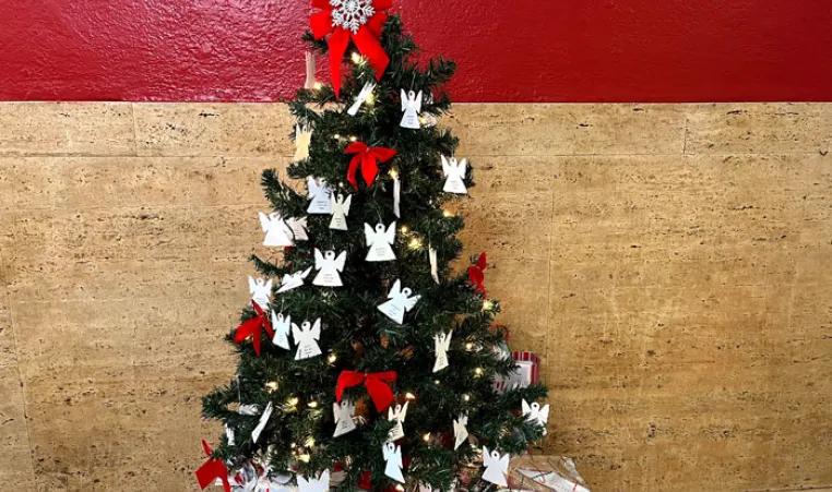 A small Christmas tree with paper angel ornaments and red bows in front of red plus tan stone wall.