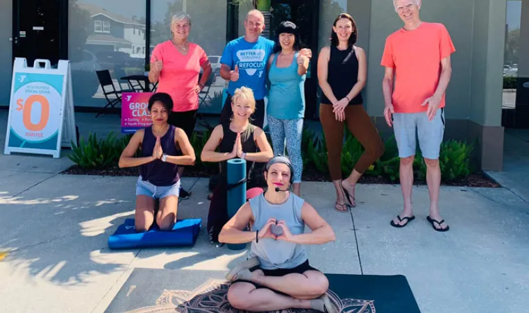 YMCA staff and members smiling outside new facility. Three people are on the ground in yoga pose with prayer hands and five others stand behind them.