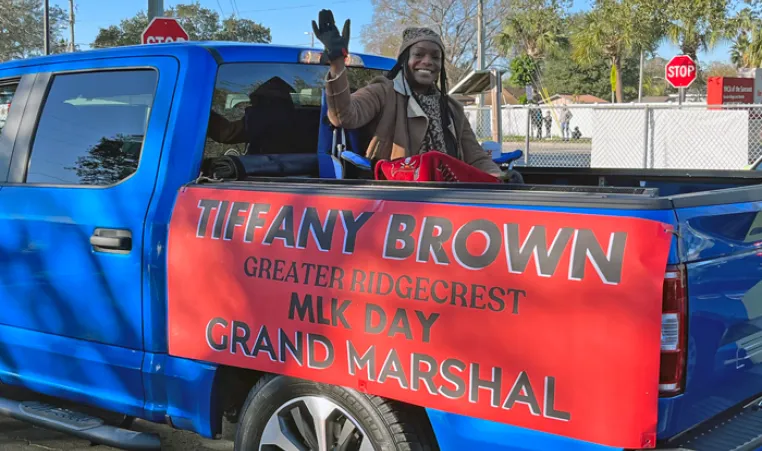 Ridgecrest leader waves from the bed of a blue truck for Greater Ridgecrest MLK Day parade. Sign is on side of truck bed, black block letters in all caps: Tiffany Brown Greater Ridgecrest MLK Grand Marshal on red background.