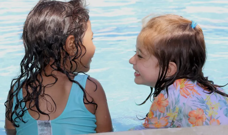 Two toddlers with wet hair and smile at each other on pool ledge during water safety class.
