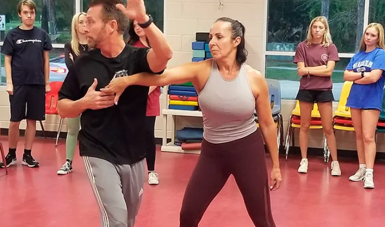 Man and woman instructors demonstrate self defense move to teens in background.