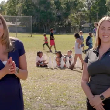 Still from video of Gayle Guyardo and Alyssa Heartstock outside in empty field, smiling. Kids playing games in after school program in the background.