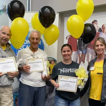 Three Greater Palm Harbor Y LIVESTRONG® at the YMCA participants and their coach at their graduation ceremony in April 2022. Balloons and poster of happy people side hugging in background.