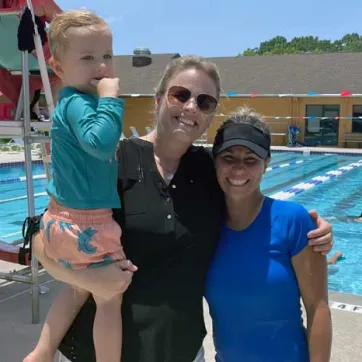 Swim instructor and mom holding young child on the outdoor pool deck. In the background, people are swimming and a lifeguard in a chair watches over them. 