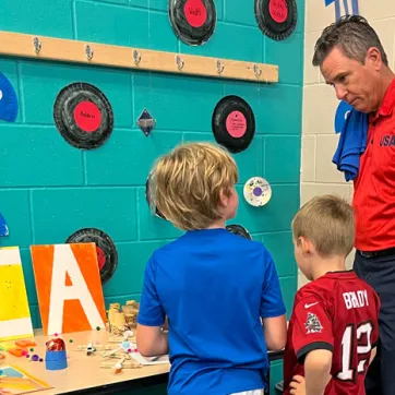 Man standing at craft display table with two children as they show off their work.