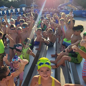 A group of 50+ youth triathletes smile while holding thumbs up on the outdoor pool deck.