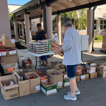 Four volunteers at James P. Gills YMCA organize food donations outside in the shade. Many brown boxes of food rest on pallets and the ground. Foreground includes a man facing away from the camera, holding cereal boxes.