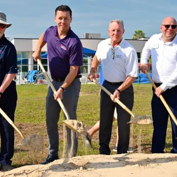 Outdoor image of four men posing for a groundbreaking photo. The subjects are holding shovels with dirt. A swimming pool with slides is in the background.
