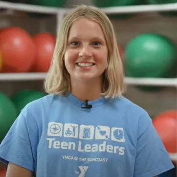 A teen girl with short blonde hair and a light blue Teen Leaders YMCA shirt smile at the camera. The background is out of focus, and shows white shelves holding green and red exercise balls.