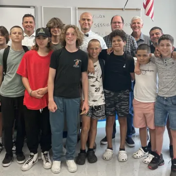 A group of five adults stand behind 10 middle school students, posing for a photo. The setting is a school classroom, a portion of the hanging American flag is shown in the background.