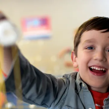 A young boy smiles as he reaches his arm out to show off the craft he made. The child's face is in focus and the craft is in the foreground, out of focus.