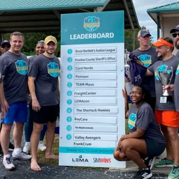 10 men and women pose by Corporate Cup Leader Board as winners for the day 