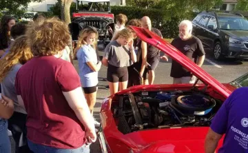 A group of teens from Teen Leaders Club at John Geigle gather around a red car with the hood lifted. Car aficionado and YMCA namesake John Geigle teaches the group about caring for cars. 