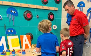 Man standing at craft display table with two children as they show off their work.