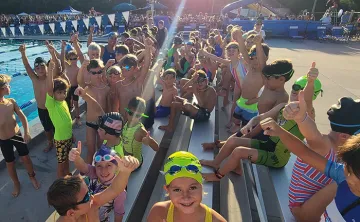 A group of 50+ youth triathletes smile while holding thumbs up on the outdoor pool deck.